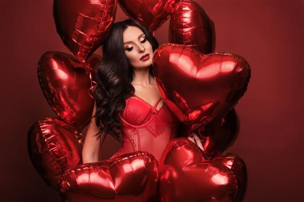 زن سکسی با لباس زیر توری قرمز و بادکنک‌هایی با قلب‌هایی که در استودیو در روز ولنتاین ژست گرفته است