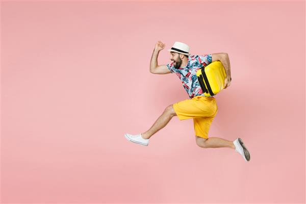 نمای کناری کامل مرد توریست مسافر شاد با کلاه تابستانی که در حال پریدن در چمدان نگهدارنده جدا شده روی پس‌زمینه صورتی است مسافر در تعطیلات آخر هفته مفهوم سفر پرواز هوایی