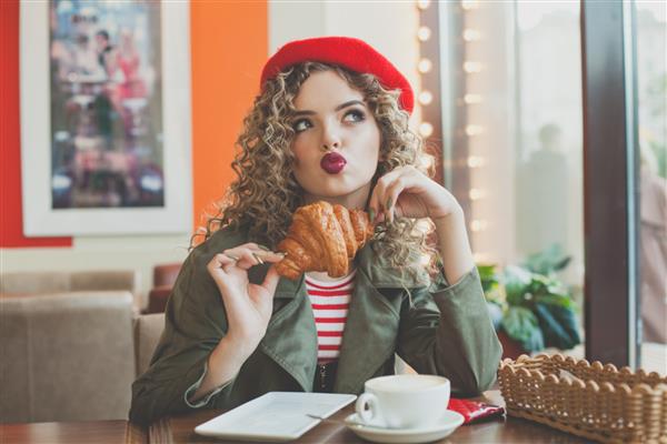 زن جوان ناز در حال نوشیدن قهوه و خوردن کروسان فرانسوی در کافه