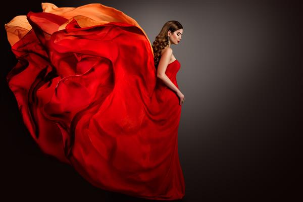 لباس قرمز زنی که روی باد پرواز می کند پرتره دختر مد زیبا در پس زمینه خاکستری مانتو متحرک