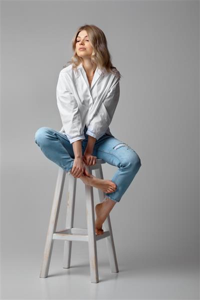 زن بلوند زیبا با موهای مجعد شلوار جین و پیراهن سفید روی صندلی در پس زمینه استودیو نشسته است
