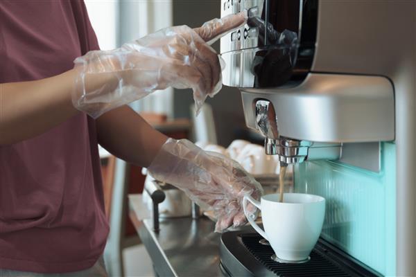 ضایعات پلاستیکی و معمولی جدید در طول کووید-19 افزایش می یابد دست های نزدیک یک زن - مهمان هتل با دستکش یکبار مصرف نوشیدنی گرم را از دستگاه قهوه خودکار در بوفه یا منطقه سلف سرویس می گیرد