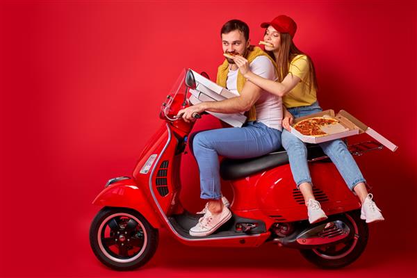 زن مو قرمز بامزه با دوست پسر پیک در حال تحویل سفارش غذا با موتور سیکلت سوار می شود زن در حال خوردن پیتزا پشت مرد نشسته است پس زمینه قرمز جدا شده