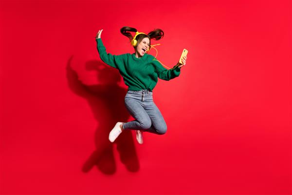 عکس تمام قد از بانوی جوان شایان ستایش پیراهن کشباف سبز پوشیده در حال گوش دادن به موسیقی در حال رقصیدن در حال پریدن پس زمینه رنگ قرمز جدا شده