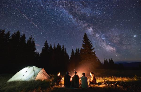 کمپینگ تابستانی عصرانه جنگل صنوبر در پس‌زمینه آسمان با ستاره‌های در حال سقوط و راه شیری گروه پنج نفره از دوستان دور هم دور آتش در کوه ها نشسته اند و از هوای تازه در نزدیکی چادر نورانی لذت می برند