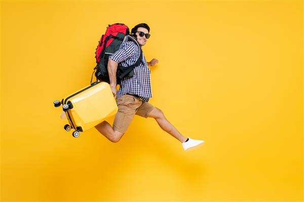 پرتره سفر گردشگر مرد جوان قفقازی هیجان‌زده با چمدانی که در هوا در پس‌زمینه زرد استودیو جدا شده می‌پرد