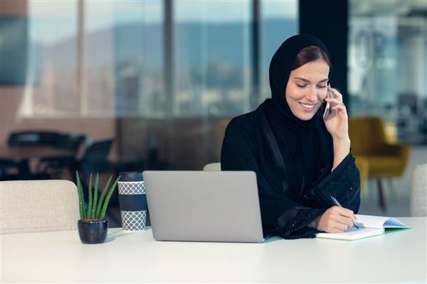 تاجر مسلمان شاد با حجاب در محل کار اداری زن عربی خندان که روی لپ تاپ کار می کند و با تلفن هوشمند صحبت می کند