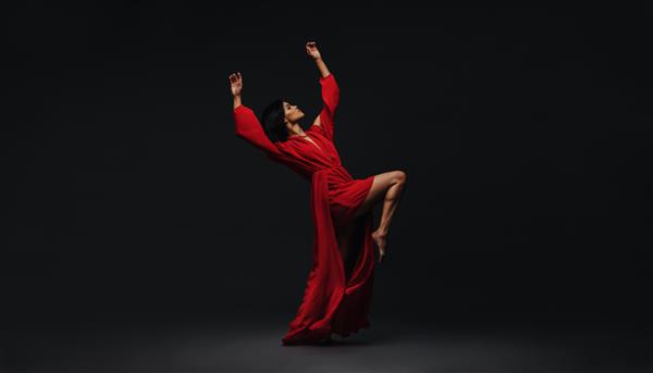 تمام طول یک زن جوان در حال رقصیدن در استودیو رقصنده زن معاصر با لباس قرمز که روی پس زمینه سیاه اجرا می کند
