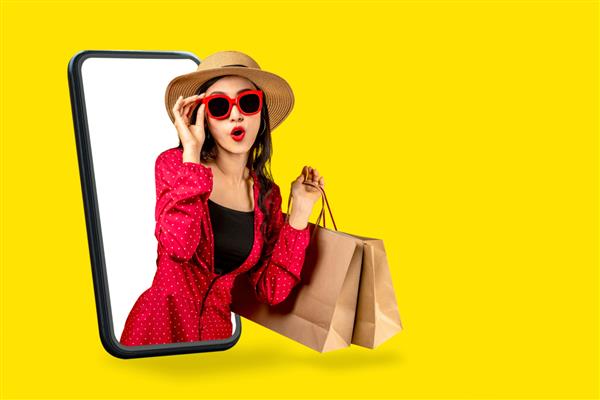 زن جوان جذاب آسیایی که کیسه خرید در دست دارد احساس هیجان شادی و شگفتی از زیبایی یا تبلیغات تخفیف فروشگاه آنلاین مد با فناوری مفهومی در تجارت الکترونیک همه‌کانالی از طریق صفحه نمایش تلفن همراه دارد