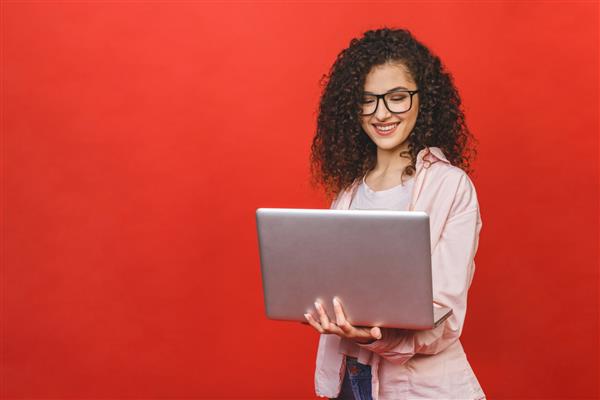 پرتره یک دختر جوان مجعد هیجان‌زده که کامپیوتر لپ‌تاپ را جدا شده روی پس‌زمینه قرمز در دست دارد