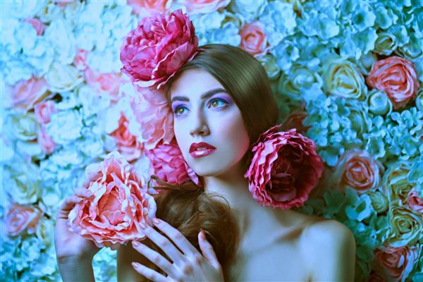 زن جوان زیبا با آرایش ملایم بنفش بنفش و گل صد تومانی در موهایش که روی پس زمینه گل رز ژست گرفته است الهام بخش بهار و تابستان مفهوم عطر لوازم آرایشی