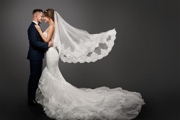 زوج عروسی عروس زیبا با لباس سفید و چادر بلند داماد زیبا در حال بوسیدن پرتره رمانتیک استودیویی