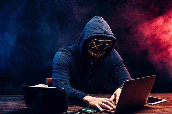 مرد جوان جنایتکار صورت خود را زیر کاپوت و ماسک پنهان می کند رمز عبور لپ تاپ را هک می کند و چیزی را تایپ می کند مرد ناشناس و ناشناس قصد هک کردن مفهوم حمله سایبری