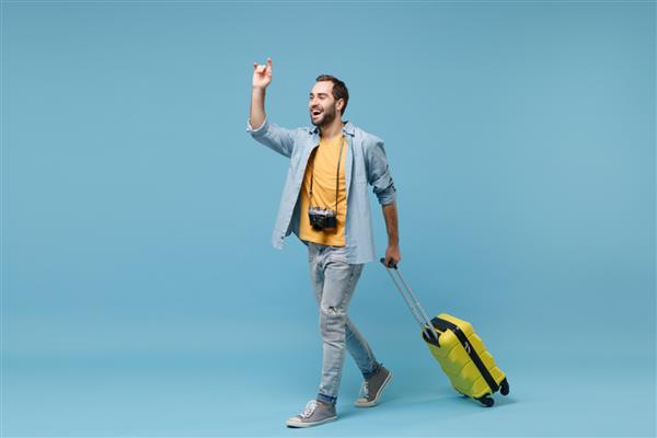 مرد گردشگر مسافر خنده دار با لباس های زرد با دوربین عکس جدا شده در پس زمینه آبی مسافر مرد در تعطیلات آخر هفته سفر پرواز هوایی چمدان را در حال تکان دادن برای تاکسی تبریک بگیرید
