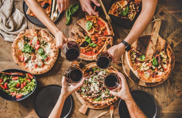 خانواده یا دوستان در حال صرف شام مهمانی پیتزا مردمانی که روی میز چوبی روستایی با انواع پیتزای ایتالیایی با نمای بالا لیوان‌ها را با شراب قرمز به هم می‌زنند ناهار فست فود جشن