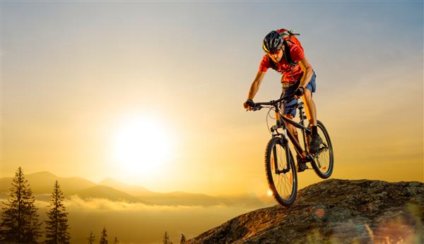 دوچرخه سوار با تی شرت قرمز در حال دوچرخه سواری در کوه های زیبا در پایین صخره در پس زمینه آسمان طلوع خورشید کانسپت دوچرخه سواری اسپرت و اندرو