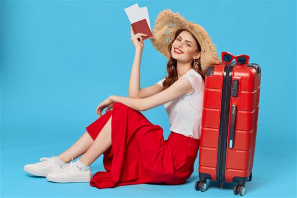 زن زیبا روی زمین نشسته و به چمدان قرمز رنگ با گذرنامه در دست و بلیط سفر تکیه داده است