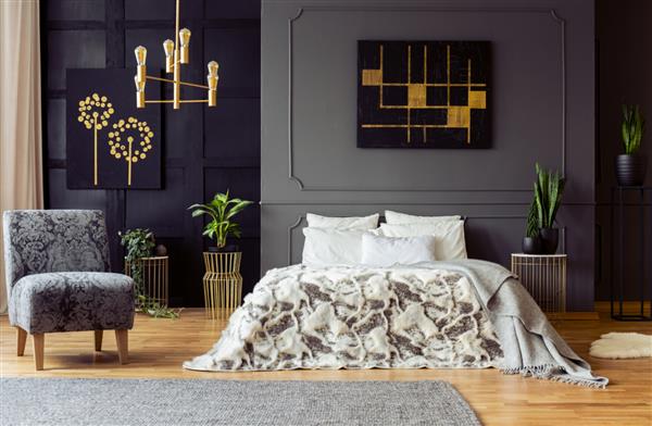 عکس واقعی از گیاهان تازه و نقاشی های سیاه و طلایی آویزان شده روی دیوار با قالب بندی در فضای داخلی اتاق خواب تاریک با تخت کینگ و صندلی راحتی گلدار