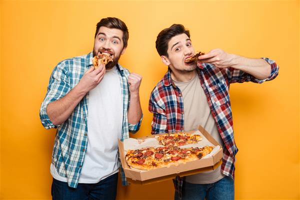 دو مرد شاد با پیراهن در حال خوردن پیتزا و نگاه کردن به دوربین در پس زمینه زرد