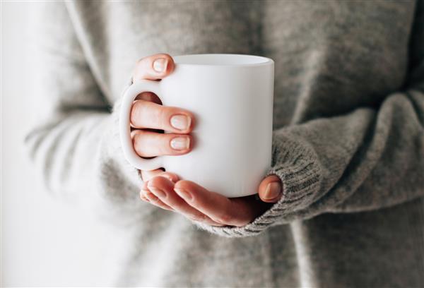 نمای نزدیک از دست های زن با یک لیوان نوشیدنی دختری زیبا با ژاکت خاکستری که در نور آفتاب صبحگاهی یک فنجان چای یا قهوه در دست دارد لیوان برای طراحی شما خالی