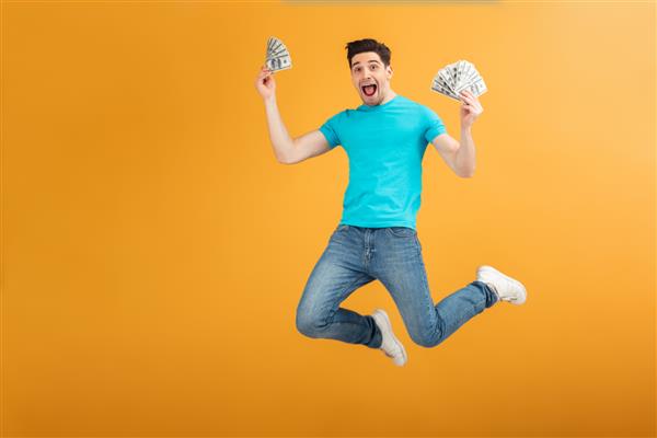 پرتره یک مرد جوان شاد با تی شرت که دسته ای از اسکناس های پول را در دست دارد و در حالی که جدا از پس زمینه زرد می پرد جشن می گیرد