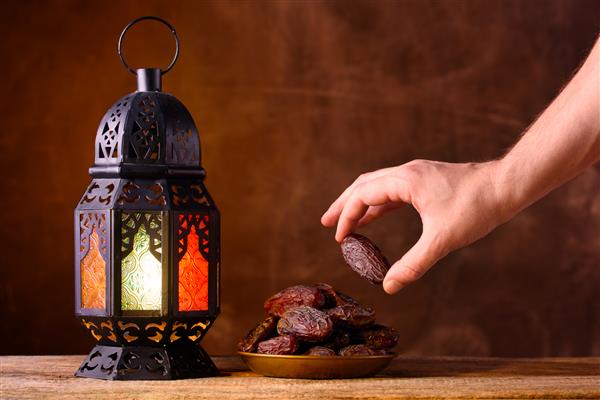 مفهوم ماه مبارک رمضان سبک زندگی مسلمان صالح روزه داری فانوس رمضان خرما دست مردی به بشقاب با خرما روی میز چوبی دراز می شود پس زمینه قهوه ای تیره