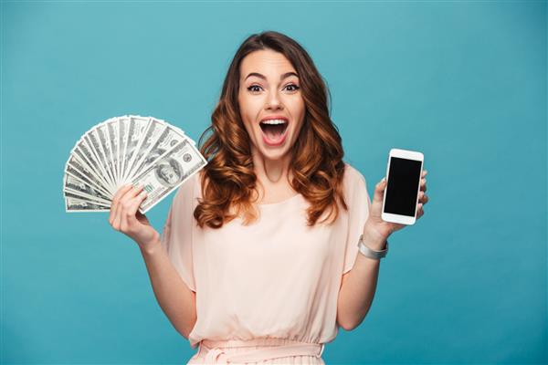 تصویر بانوی جوان هیجان زده ایزوله شده روی پس زمینه آبی دوربینی که نمایشگر تلفن همراهی را نشان می‌دهد که پول در دست دارد