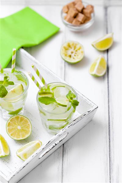 کوکتل کایپیرینیا یا موهیتو با لیمو شکر قهوه‌ای یخ و برگ‌های نعنا در لیوان‌های زیبا و برش مرکبات سبز روی زمینه چوبی سفید نوشیدنی الکلی تابستانی