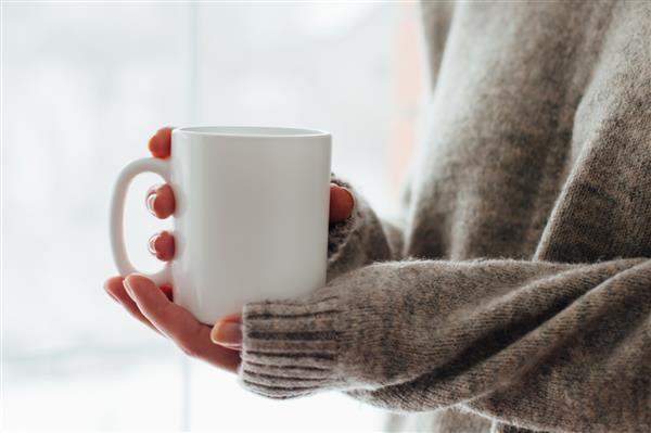 دست‌های زنانه از نزدیک که لیوان سفید در دست دارند با فضای خالی کپی برای پیامک تبلیغاتی یا محتوای تبلیغاتی قهوه یا چای شیرین