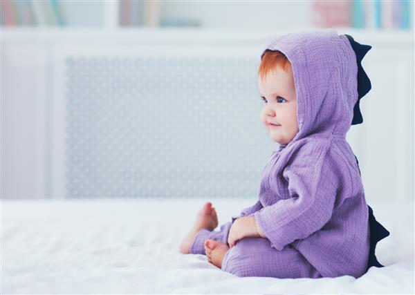 خندان زیبا نه ماهه نوزاد دختر با لباس دایناسور روی تخت نشسته است