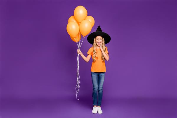 عکس در اندازه کامل از مهمانی هالووین خانم کوچولوی هیجان‌زده که بادکنک‌های هوا را در دست گرفته است برای مهمانی با تم بچه‌ها می‌پوشد تی‌شرت نارنجی کلاه جادوگر با پس‌زمینه بنفش رنگ جدا شده