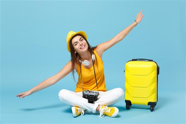 زن توریست مسافر با کلاه لباس غیررسمی زرد با دوربین عکس چمدان جدا شده در پس‌زمینه آبی مسافر زن در حال سفر به خارج از کشور برای سفر در تعطیلات آخر هفته مفهوم سفر پرواز هوایی