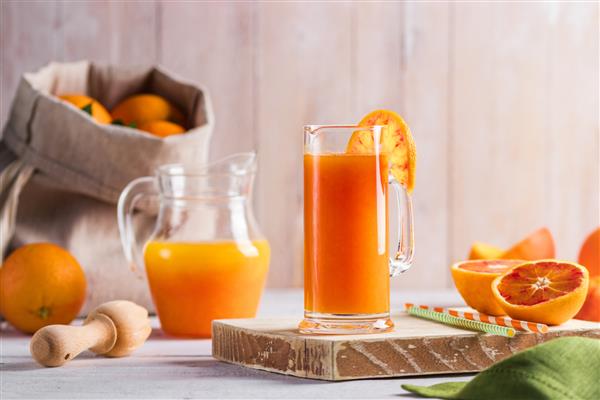 یک لیوان آب پرتقال تازه فشرده و پرتقال خونی روی میز چوبی حال و هوای تابستانی روشن