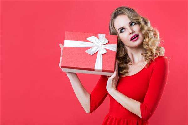 زن متعجب جعبه هدیه قرمز را در لباس قرمز نگه می دارد دختر زیبا با موهای مجعد