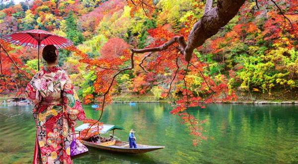 دختر ژاپنی لباس سنتی کیمونو می پوشد و در پارک آراشیاما با پس زمینه پاییزی شهر کیوتو ژاپن ایستاده است