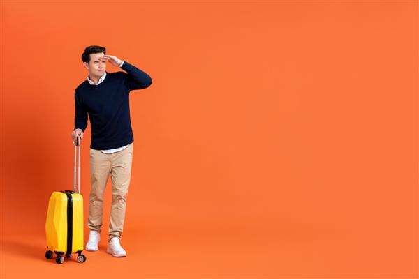 مرد جوان خوش‌تیپ جهانگرد آمریکایی که چمدان‌های آماده سفر را در دست گرفته و با دست روی پیشانی جدا شده روی پس‌زمینه نارنجی با فضای کپی به دور نگاه می‌کند