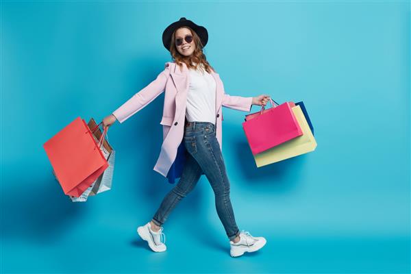 خرید به عنوان نوعی فعالیت اوقات فراغت و کاهش دهنده استرس موثر است خانمی شاد با کلاه مشکی عینک آفتابی کت صورتی شلوار جین و کفش کتانی با کیسه‌های خرید رنگارنگ در دو دست راه می‌رود