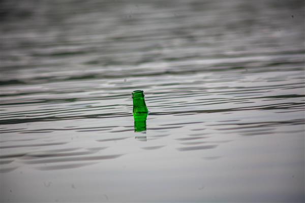 بطری شیشه‌ای سبز شناور در آب دریاچه با موج‌های آرام عکس باکیفیت آلودگی محیط زیست و سطح آب