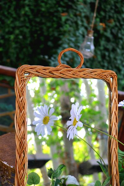 آینه تزئینی با قاب حصیری و دستبافت که گل‌های بابونه در آن منعکس شده‌اند