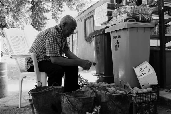 تصویر سیاه و سفید از پیرمرد دستفروش با میوه‌های حیاطی در کنار سطل‌های زباله در روستا