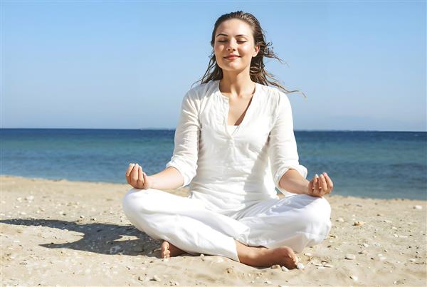 زن در حال مدیتیشن و یوگا در ساحل دریا با لباس سفید ویژه پوستر دیواری سالن‌های یوگا، کلینیک‌های زیبایی و دکوراسیون اتاق استراحت