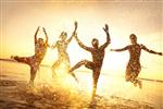 گروهی از جوانان شاد در حال رقصیدن و سمپاشی در ساحل در غروب زیبای تابستان
