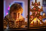 پسر ناز خندان که هنگام کریسمس کنار پنجره ایستاده است با چراغ های رنگارنگ از درخت کریسمس در پس زمینه فوکوس انتخابی