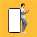 برنامه عالی مرد هیجان زده تکیه بر تلفن هوشمند بزرگ با صفحه نمایش سفید خالی و استفاده از تلفن همراهش پسر شادی که در شبکه های اجتماعی چت می کند ایستاده روی پس زمینه زرد تصویر ماکت تمام طول بدن